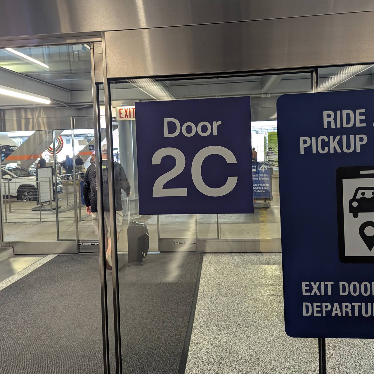 Sign indicating Door 2C in an airport terminal, with a Ride Pickup sign nearby and passengers with luggage beyond the glass doors.