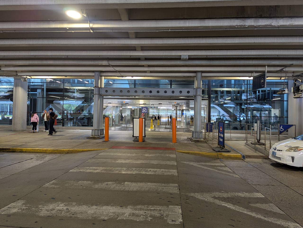 Airport terminal entrance with glass doors, orange bollards, and people waiting near a crosswalk under an overpass.