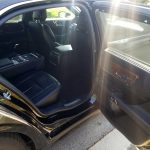 Open rear door view of a black luxury SUV interior with black leather seats and wood trim sunlit by outside light.