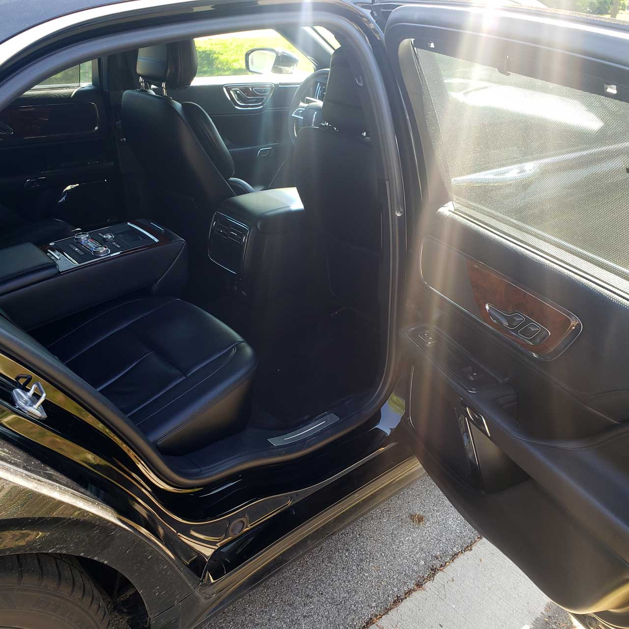 Open rear door view of a black luxury SUV interior with black leather seats and wood trim sunlit by outside light.