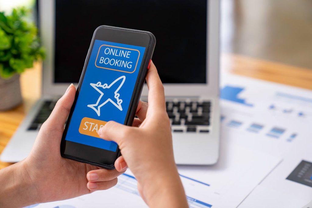 Hand holding a smartphone displaying an online flight booking screen with an airplane icon and Start button on a desk surface