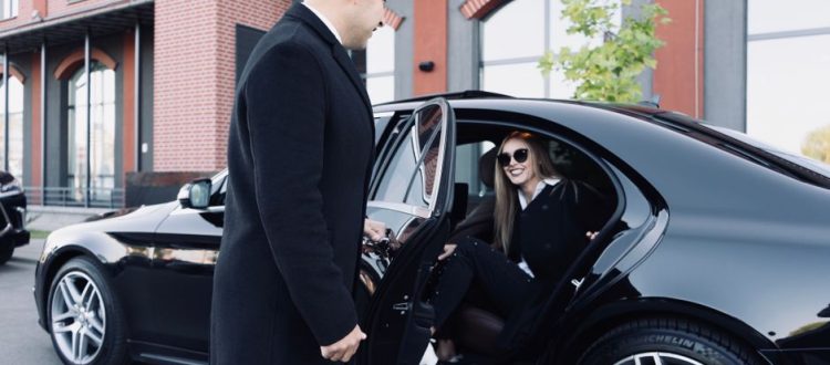 Man in a dark overcoat helps a woman into a sleek black car outside a brick building.