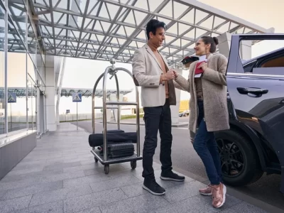 Two travelers with a luggage trolley beside a dark vehicle and glass terminal exterior