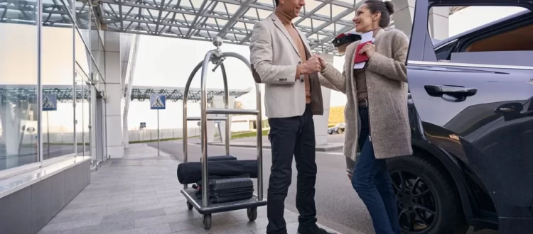 Two travelers with a luggage trolley beside a dark vehicle and glass terminal exterior