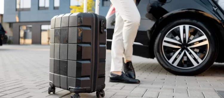 A rolling suitcase beside a traveler in light trousers and a black car outside a building