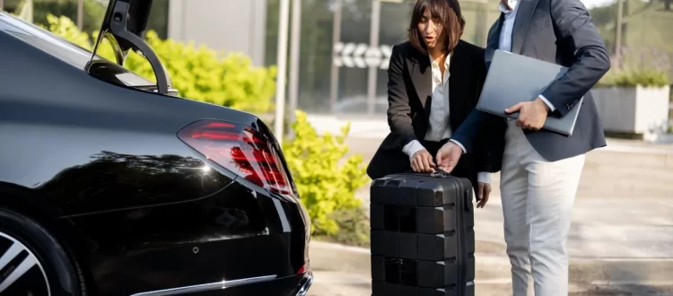 Two colleagues with a suitcase and laptop beside the open trunk of a black sedan