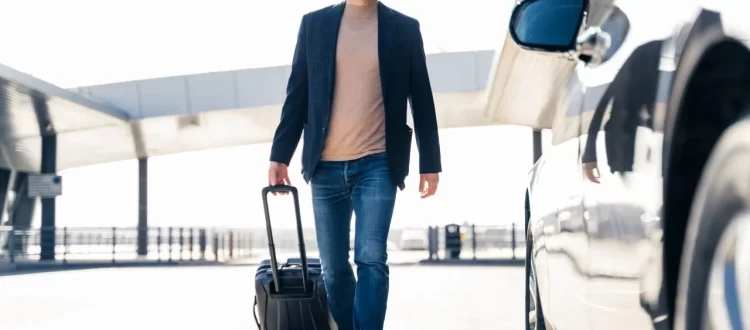 A masked man with a rolling suitcase walking beside a silver car outside a terminal