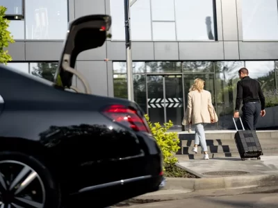 Two travelers with a suitcase approaching a glass building beside a dark sedan