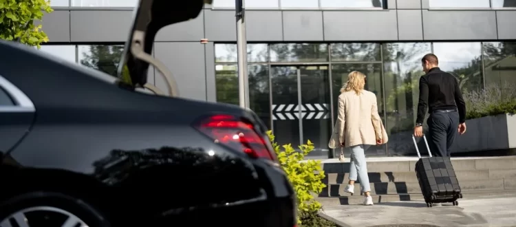 Two travelers with a suitcase approaching a glass building beside a dark sedan