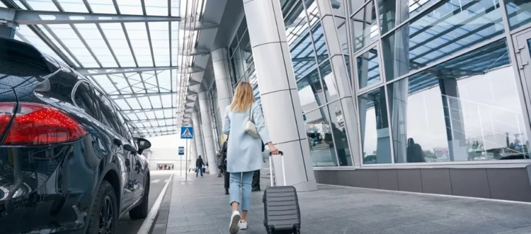 A woman in a light blue suit pulling a gray suitcase beside a black car and glass terminal