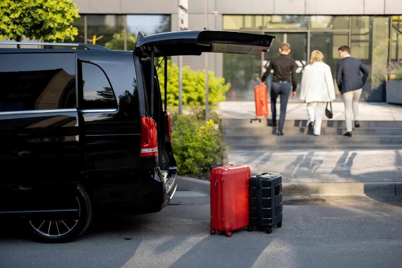 Suitcases beside an open black van while three adults walk toward a building entrance