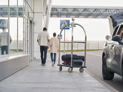 Two travelers beside glass doors, a luggage trolley and a dark vehicle