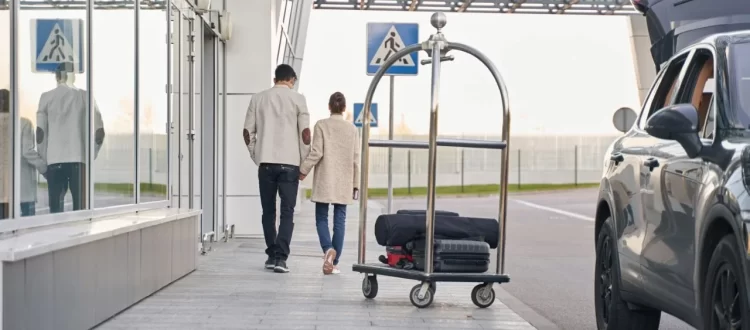 Two travelers beside glass doors, a luggage trolley and a dark vehicle