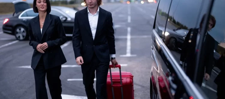 Two business travelers with a red rolling suitcase beside a dark passenger van