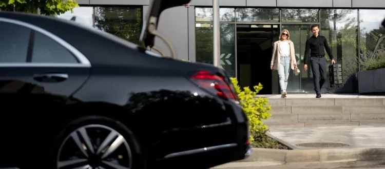 Two adults near the open trunk of a black sedan outside a glass-fronted building