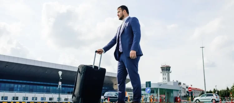 A man in a blue suit pulling a black suitcase outside an airport terminal