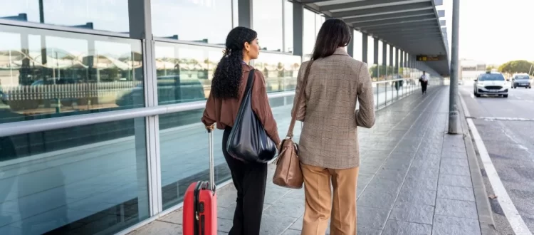 Two travelers with bags and a rolling suitcase walking beside a glass terminal exterior